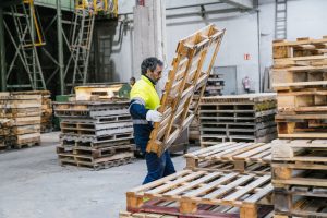 Factory worker dressed in high visibility clothing, carrying a wooden pallet through a bustling recycling plant during daylight hours
