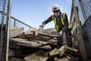 Young man wearing hard hat and reflective vest standing on top of stack of recycled wood.